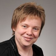 Lächelnde Person mit kurzem, blonden Haar im Porträt Die Frau hat ein offenes, freundliches Lächeln und trägt ein schwarzes Kleidungsstück, während sie in die Kamera schaut.