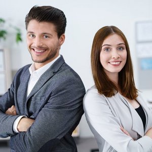 In einem hellen Büro posieren ein Mann und eine Frau, beide mit freundlichen Gesichtsausdrücken und professioneller Kleidung.